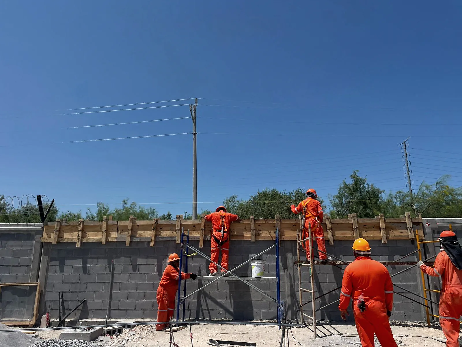 Cuadrilla de overol naranja arma, desde un andamio tubular y un tablón apoyado en una escalera de madera, la cimbra de la cadena de cerramiento de la barda perimetral de block, en la Estación de Regulación y Medición Anzalduas