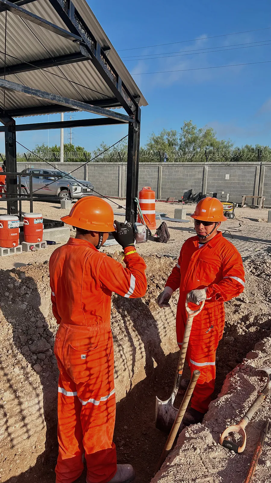 Dos trabajadores de overol naranja y casco excavan una zanja a pala junto al cobertizo de la Estación Anzalduas